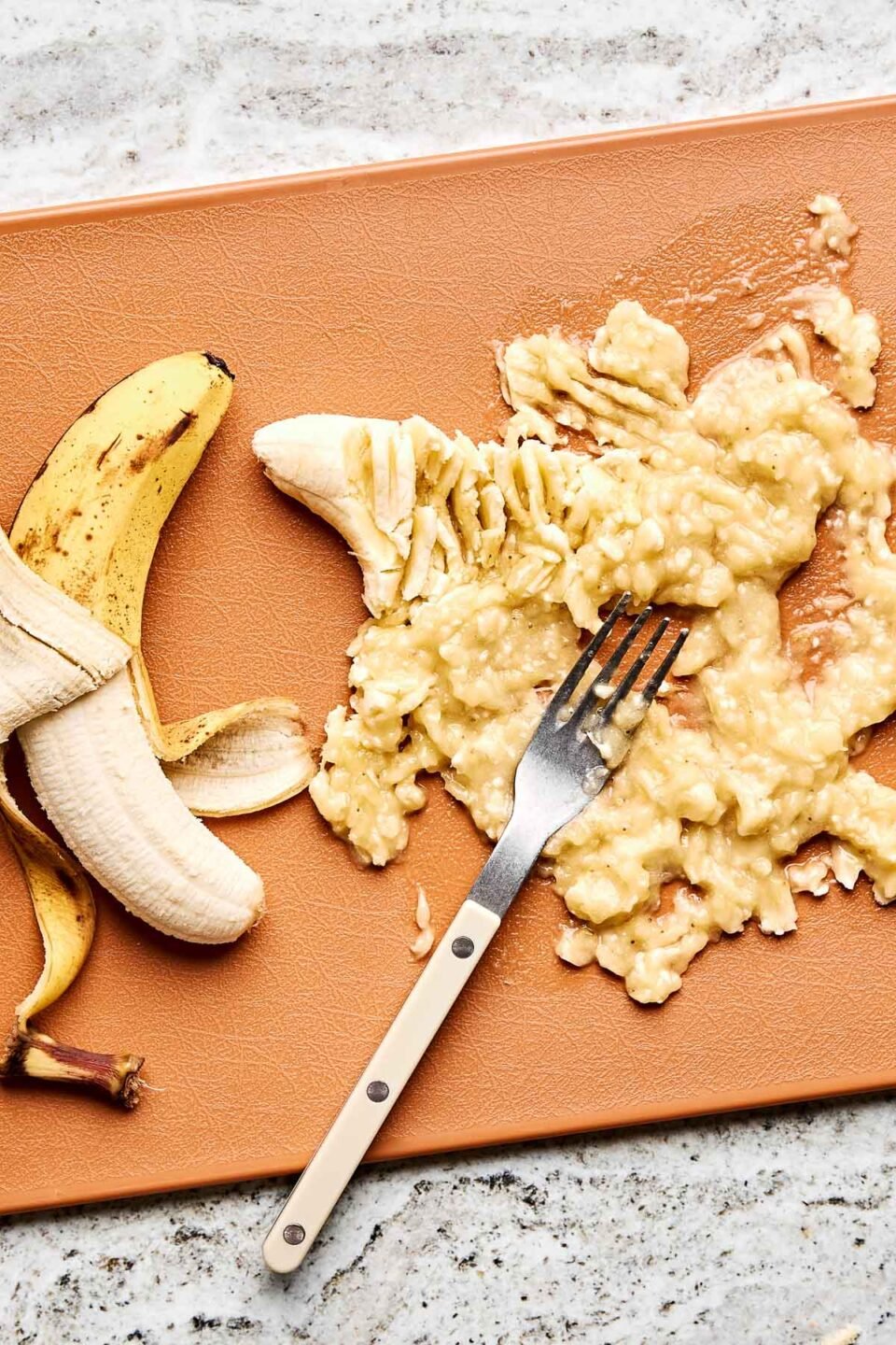 An overhead shot of a half-peeled banana and mashed banana on a light brown cutting board atop a grey textured surface.