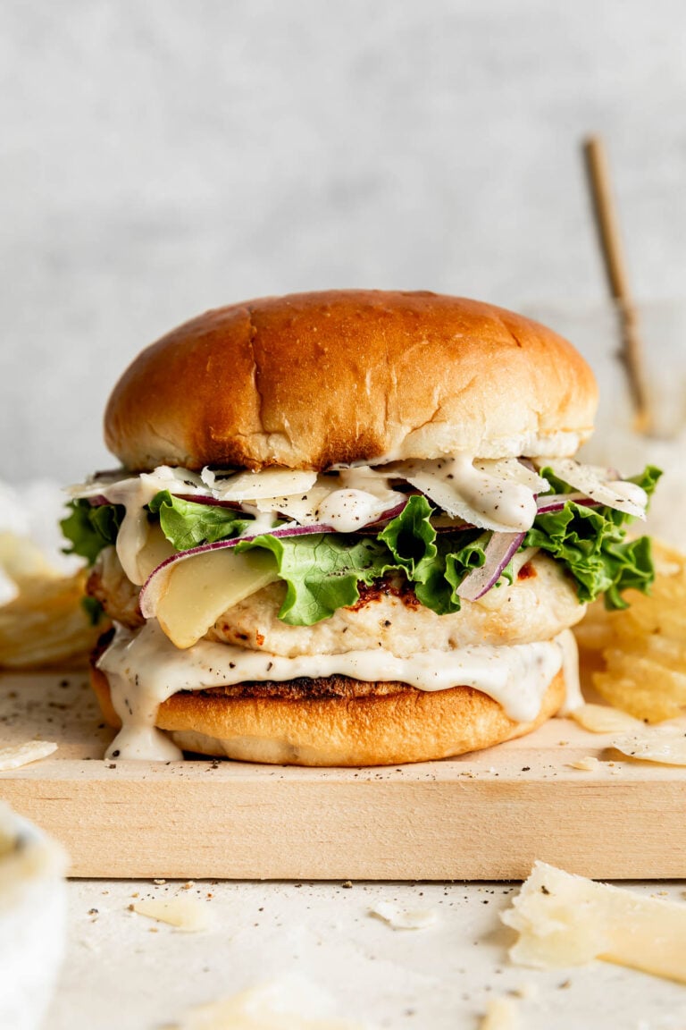 A side shot of an assembled chicken Caesar burger on a bun, topped with Caesar dressing, lettuce, cheese and red onions. The burger is sitting on a wooden board next to ridged potato chips on an off-white surface.