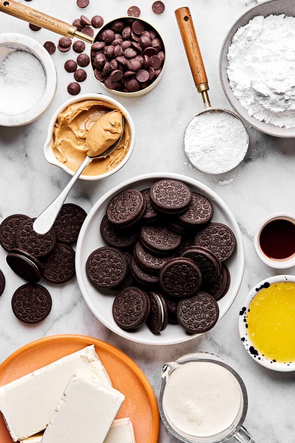 An overhead shot of ingredients displayed on a white marble surface: Oreo cookies, peanut butter, chocolate chips, cream cheese, kosher salt, powdered sugar, vanilla extract, and heavy cream.
