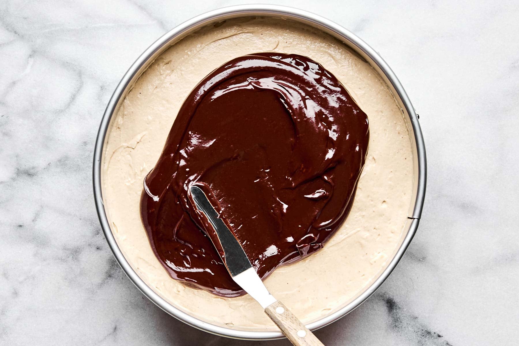 An overhead shot of chocolate ganache being spread over cheesecake batter in a springform pan atop a white marbled surface.