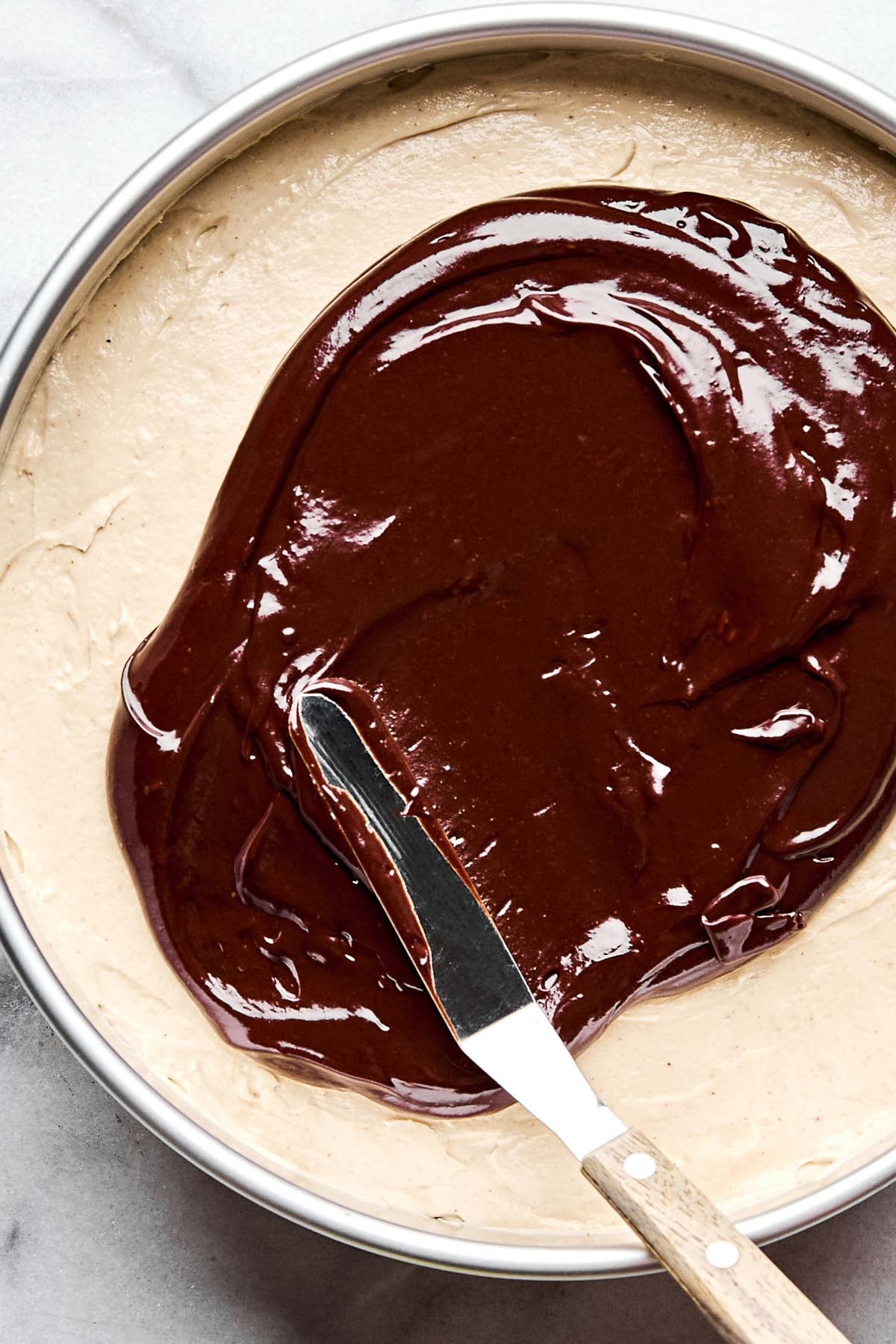 An overhead shot of chocolate ganache being spread over cheesecake batter in a springform pan atop a white marbled surface.