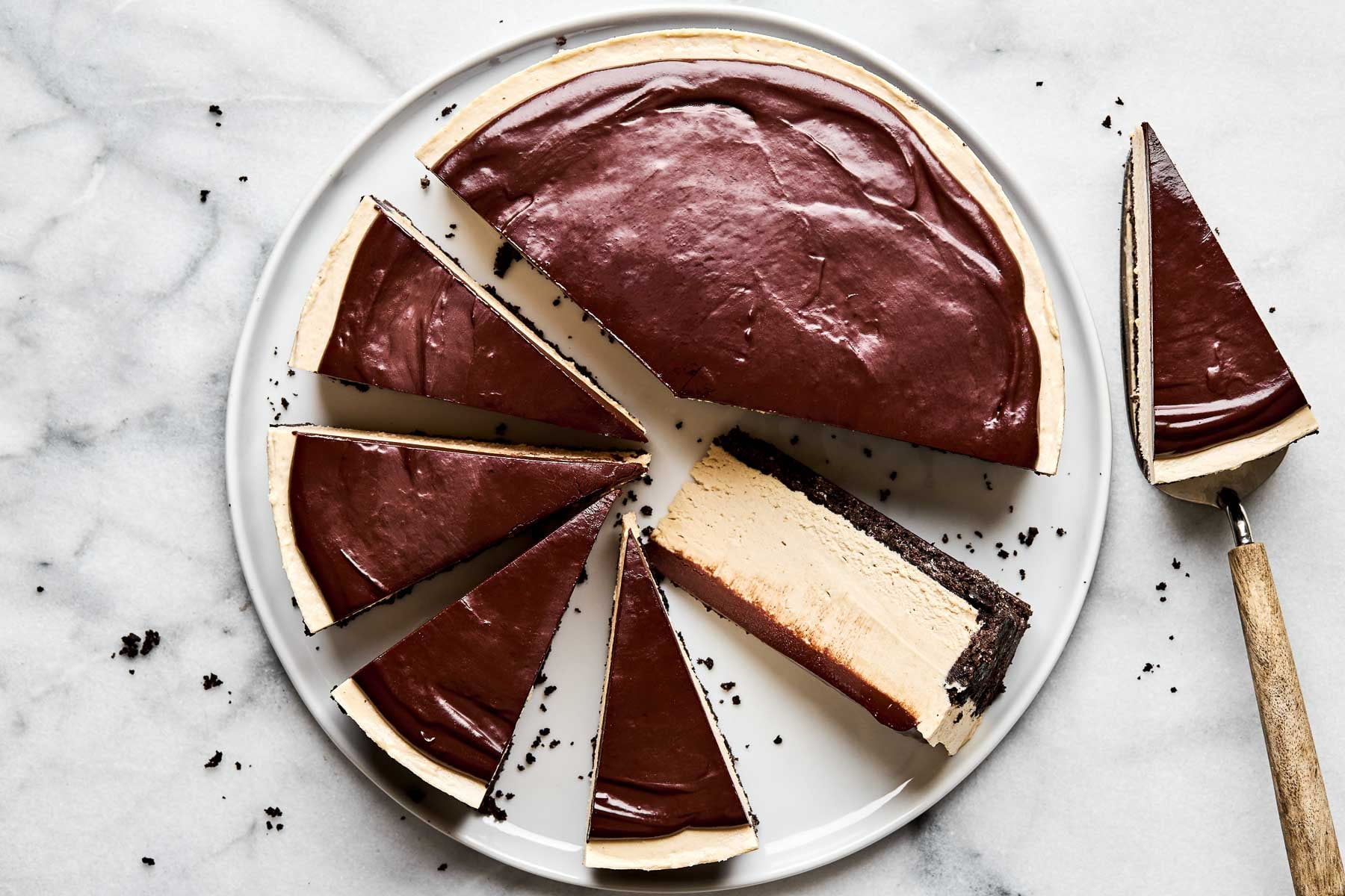 An overhead shot of a partially sliced peanut butter cheesecake on a large white plate atop a white marbled surface. One slice has been turned on its side to show the cheesecake filling, and another slice rests on a pie server.