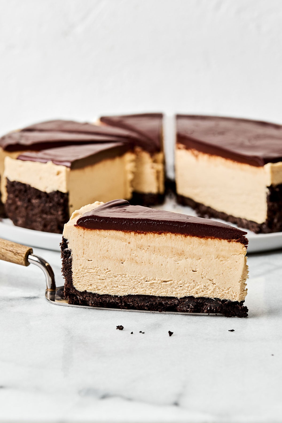 A side angle shot of a slice of peanut butter cheesecake on a pie server on a white marbled surface. A partially sliced cheesecake sits on a plate in the background.