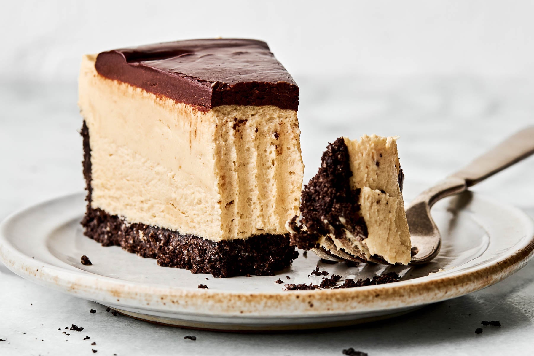 A close-up side shot of a slice of peanut butter cheesecake. A fork has taken a bite from the front of the slice, and the bite rests on the fork on a small white plate atop a white surface.