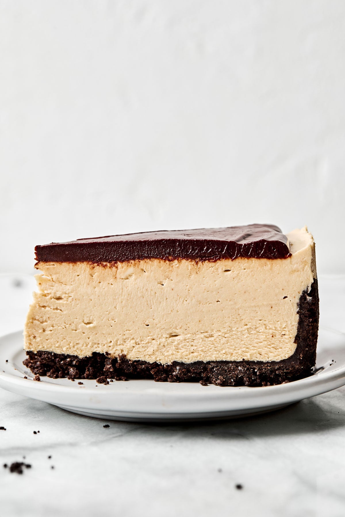 A side shot of a slice of peanut butter cheesecake on a small white plate atop a white marbled surface.