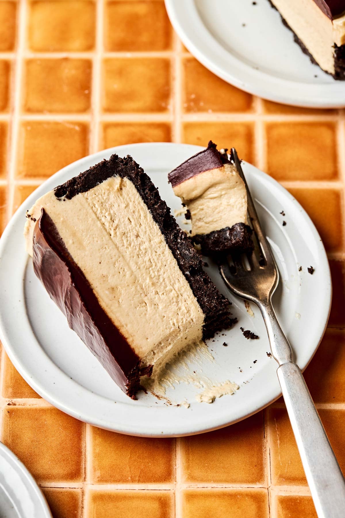 An overhead shot of a slice of peanut butter cheesecake on a small white plate, with a bite taken out and resting on a fork. The plate sits on an orange tiled surface.