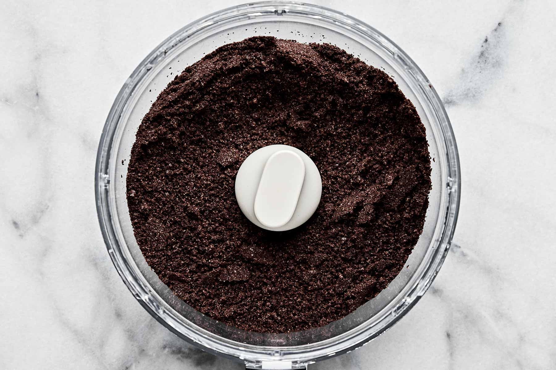 An overhead shot of crumbled Oreo cookies in the bowl of a food processor atop a white marbled surface.