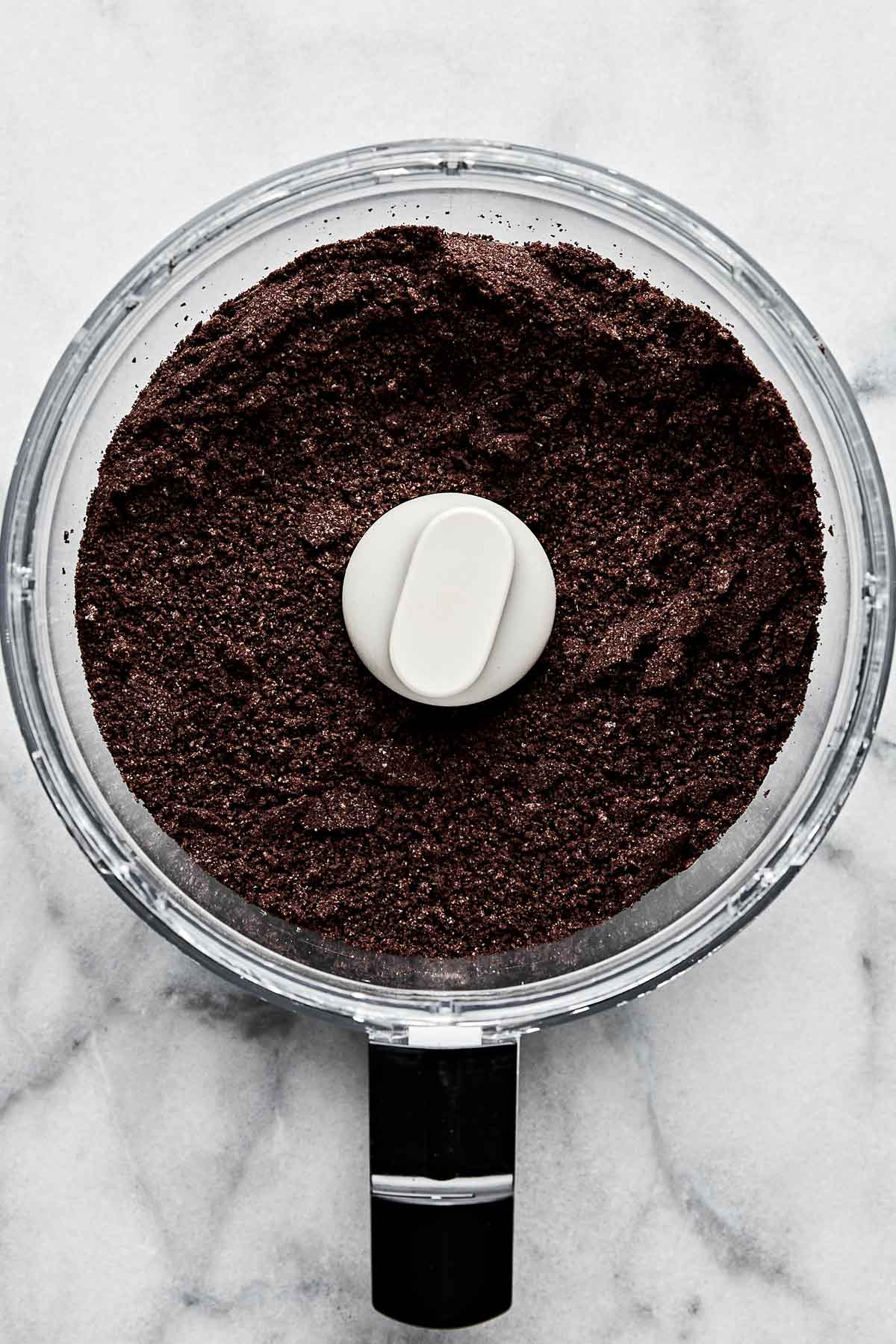An overhead shot of crumbled Oreo cookies in the bowl of a food processor atop a white marbled surface.