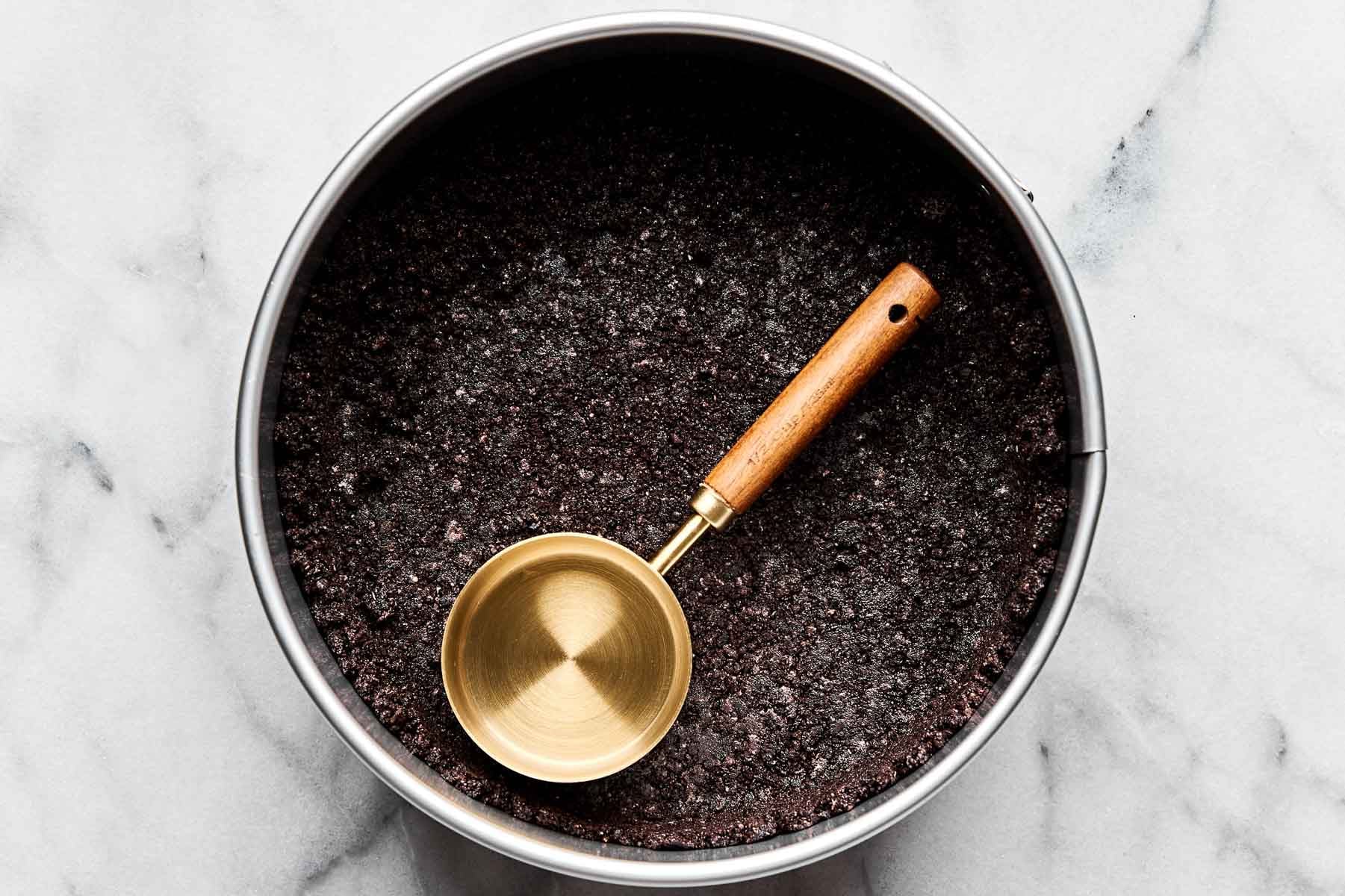 An overhead shot of Oreo crust pressed into the bottom and edges of a springform pan atop a white marbled surface. A gold, wood-handled measuring cup rests in the pan.