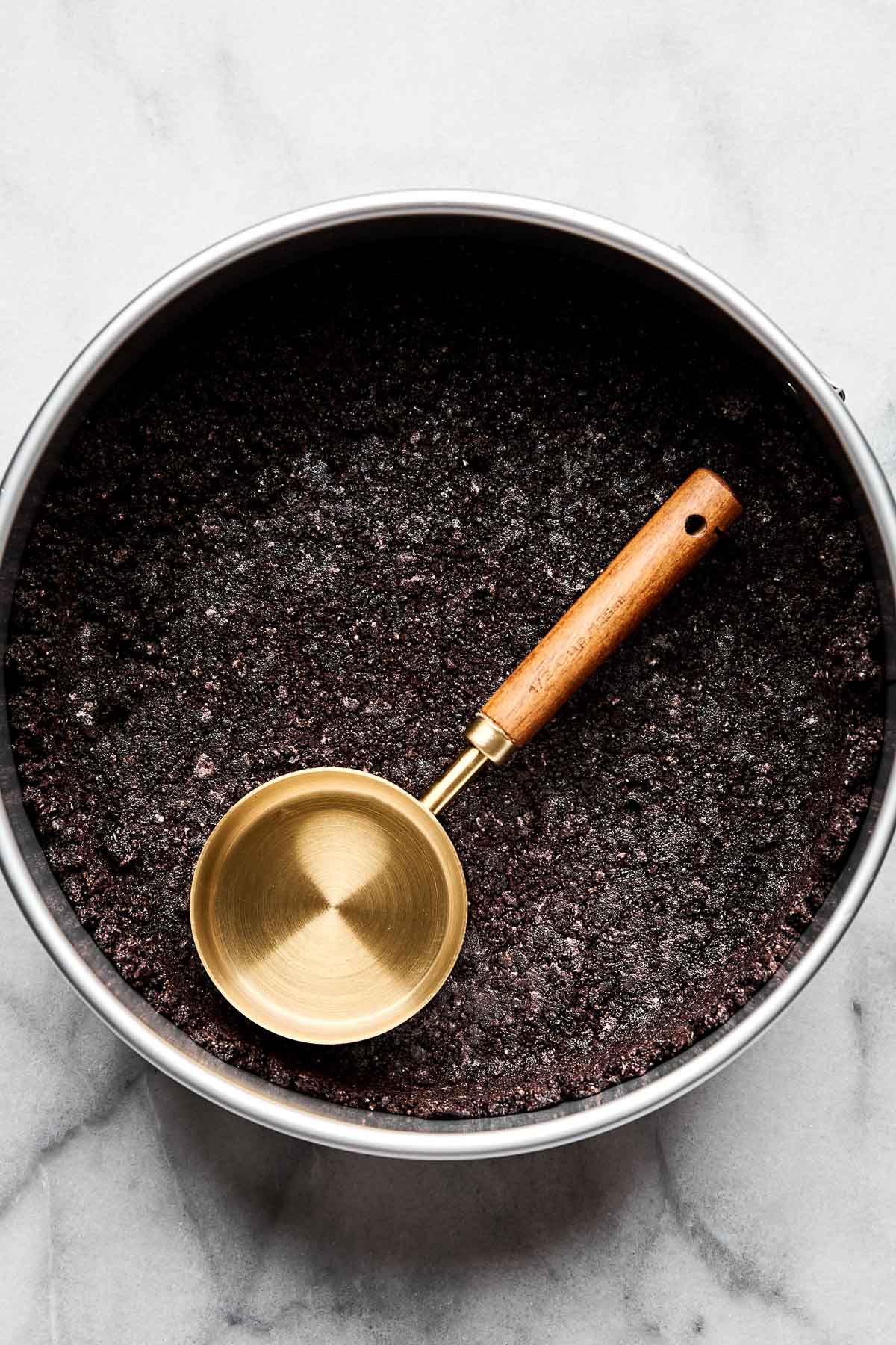An overhead shot of Oreo crust pressed into the bottom and edges of a springform pan atop a white marbled surface. A gold, wood-handled measuring cup rests in the pan.