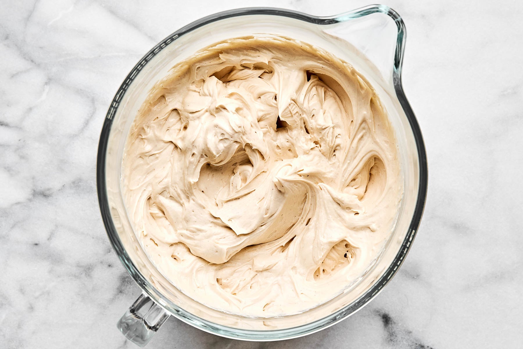 An overhead shot of cheesecake batter in a glass batter bowl atop a white marbled surface.