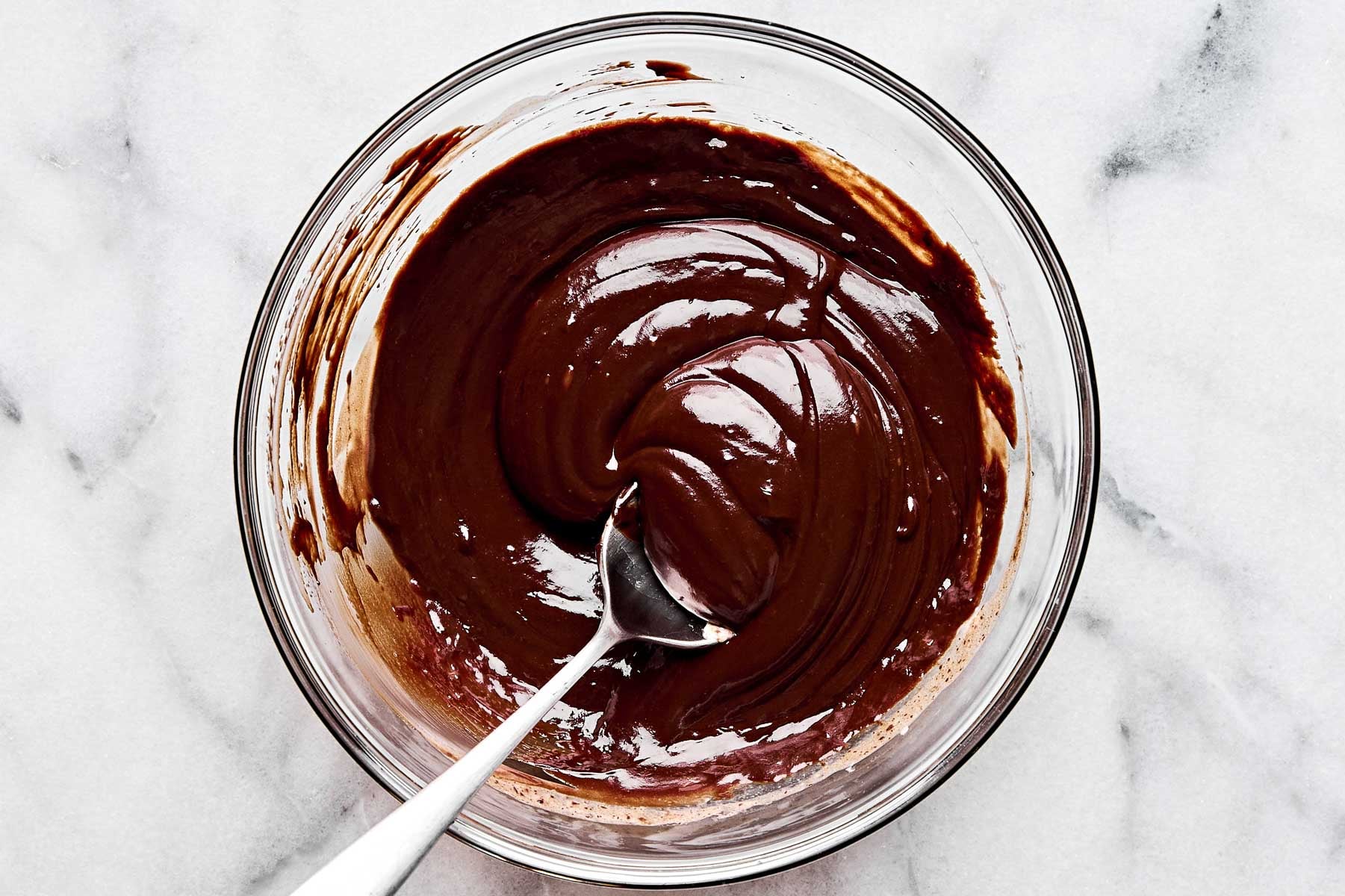An overhead shot of chocolate ganache in a glass bowl with a spoon atop a white marbled surface.