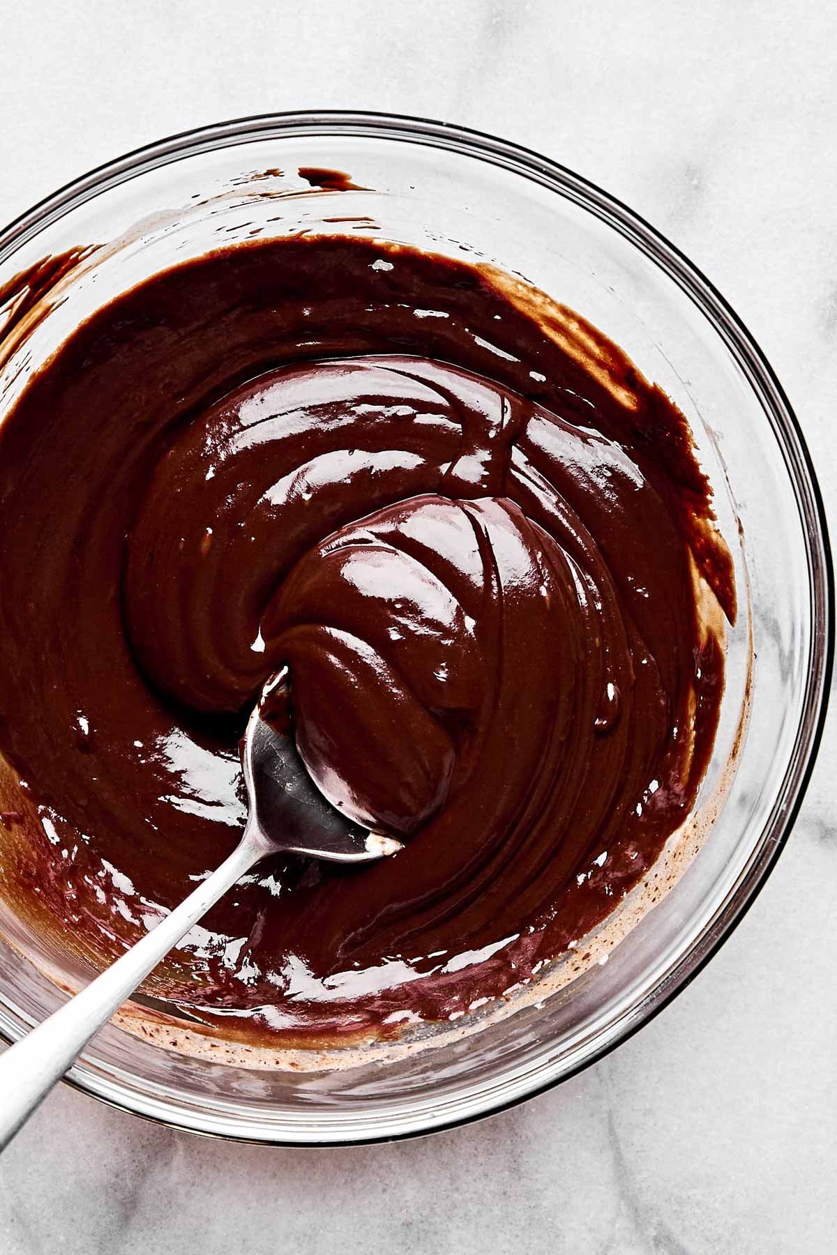 An overhead shot of chocolate ganache in a glass bowl with a spoon atop a white marbled surface.