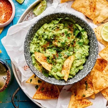 An overhead shot of a small bowl of guacamole surrounded by tortilla chips on a parchment-lined plate atop a blue tiled surface. The plate is surrounded by lime wedges and halves and a small dish of tajin.