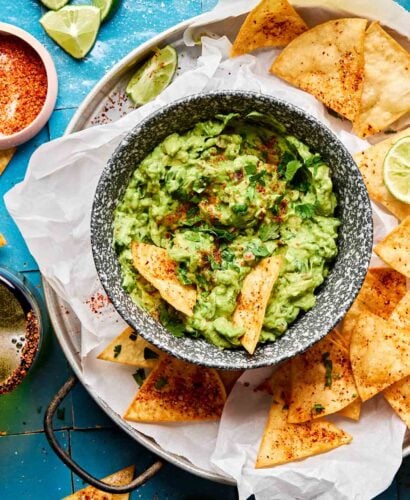 An overhead shot of a small bowl of guacamole surrounded by tortilla chips on a parchment-lined plate atop a blue tiled surface. The plate is surrounded by lime wedges and halves and a small dish of tajin.