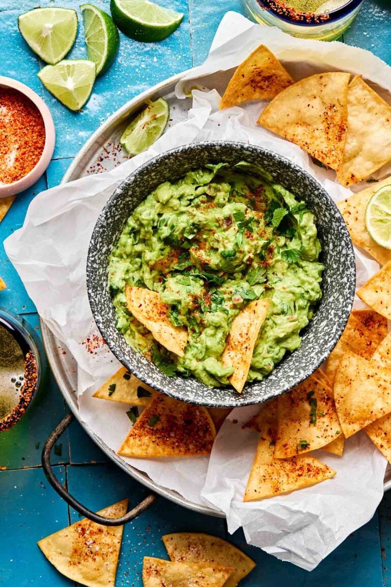 An overhead shot of a small bowl of guacamole surrounded by tortilla chips on a parchment-lined plate atop a blue tiled surface. The plate is surrounded by lime wedges and halves and a small dish of tajin.