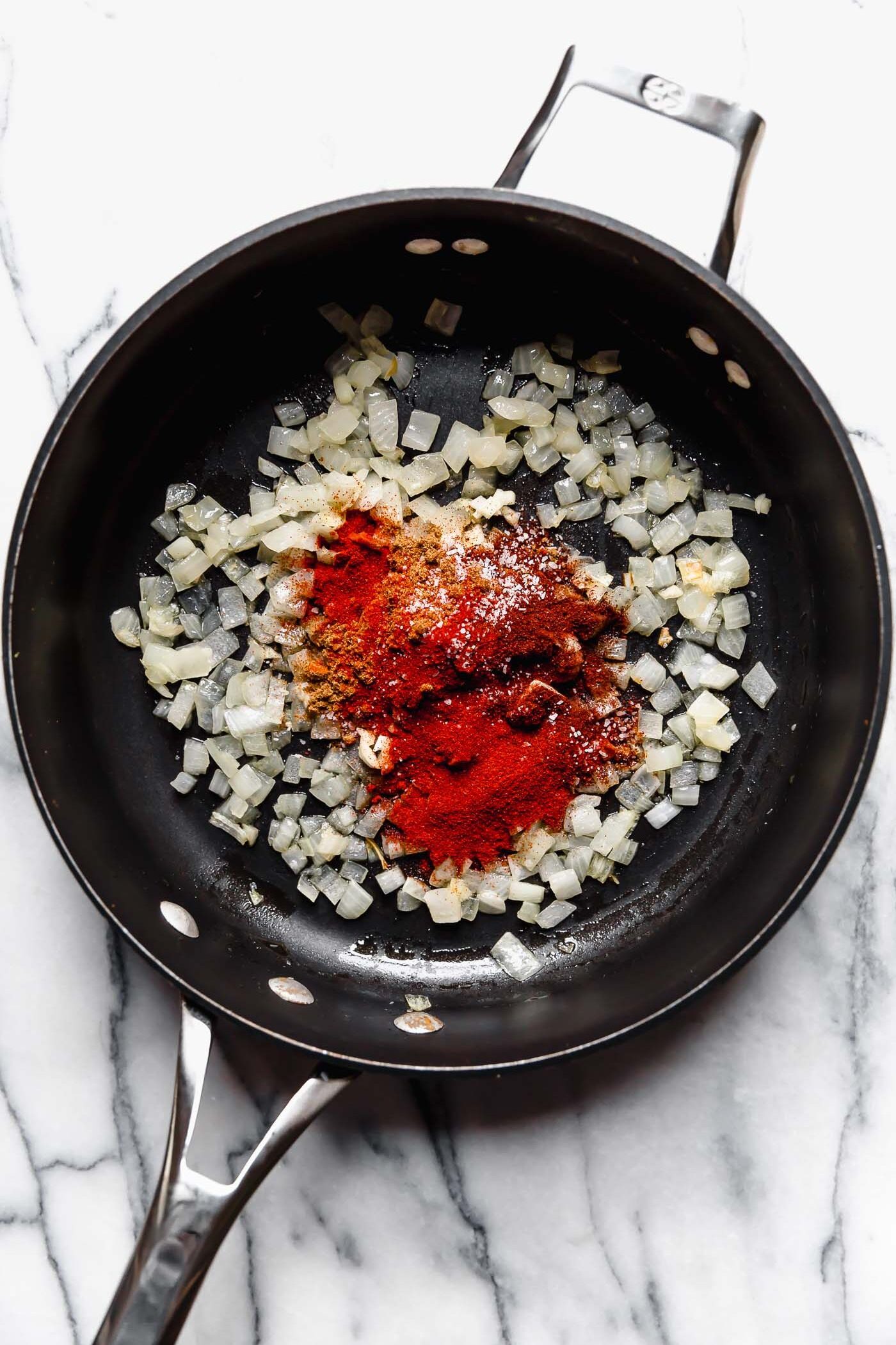 Softened onion shown in a black skillet, topped with a taco spice blend. The skillet sits atop a white marble surface.