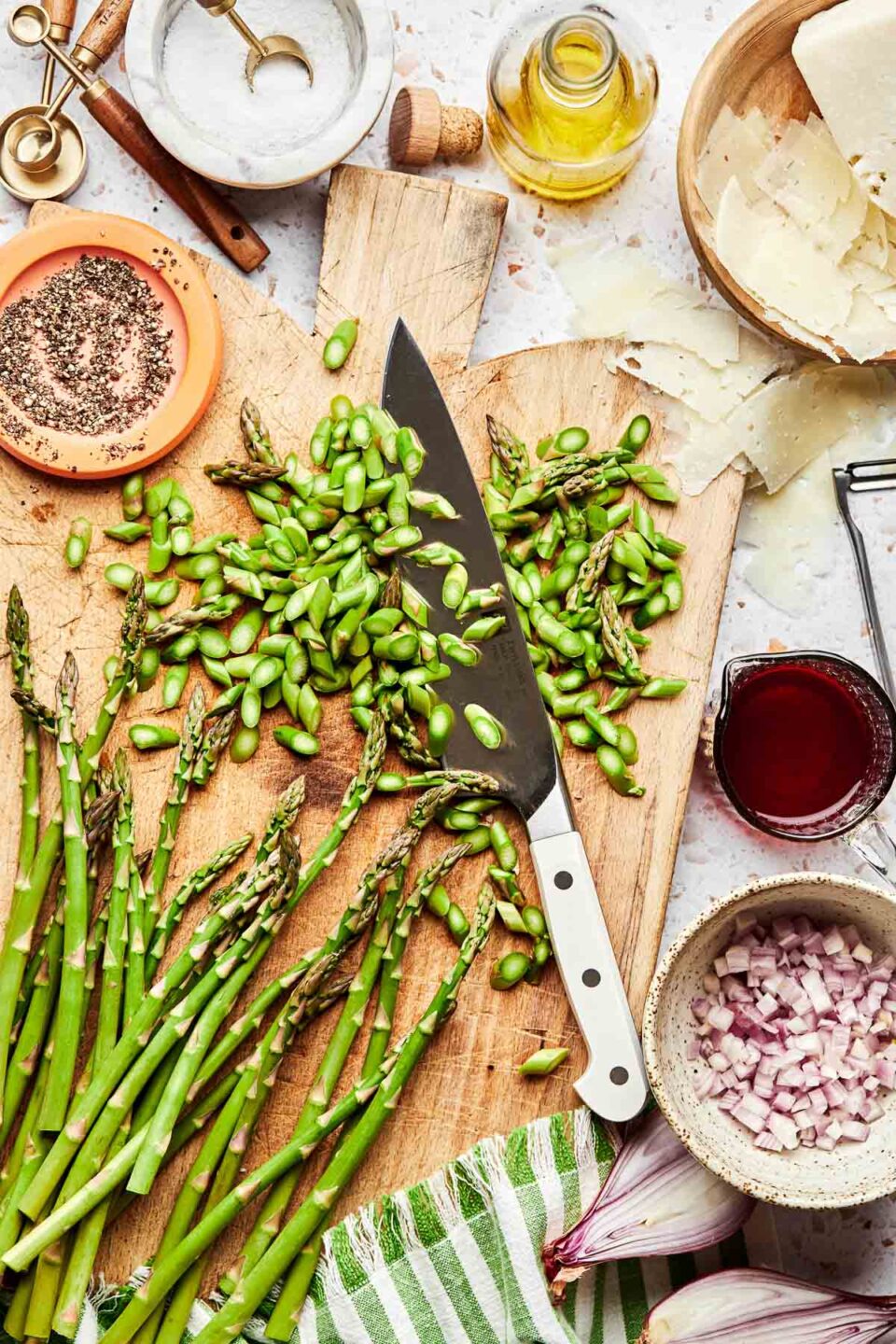 An overhead shot of ingredients displayed on a wooden board and a white textured surface: whole and chopped asparagus, shallots, pecorino cheese, red wine vinegar, olive oil, salt and pepper.