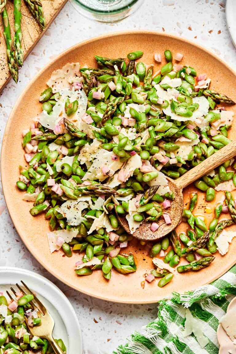 An overhead shot of raw asparagus salad on a large brown serving plate with a wooden serving spoon. The plate sits on a white textured surface, surrounded by a green and white striped towel, a small plate of salad, and asparagus spears on a wooden board.