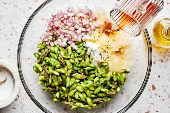 An overhead shot of red wine vinegar being poured over a large glass bowl of chopped asparagus, shallots, pecorino romano, olive oil, salt and pepper on a white textured surface.