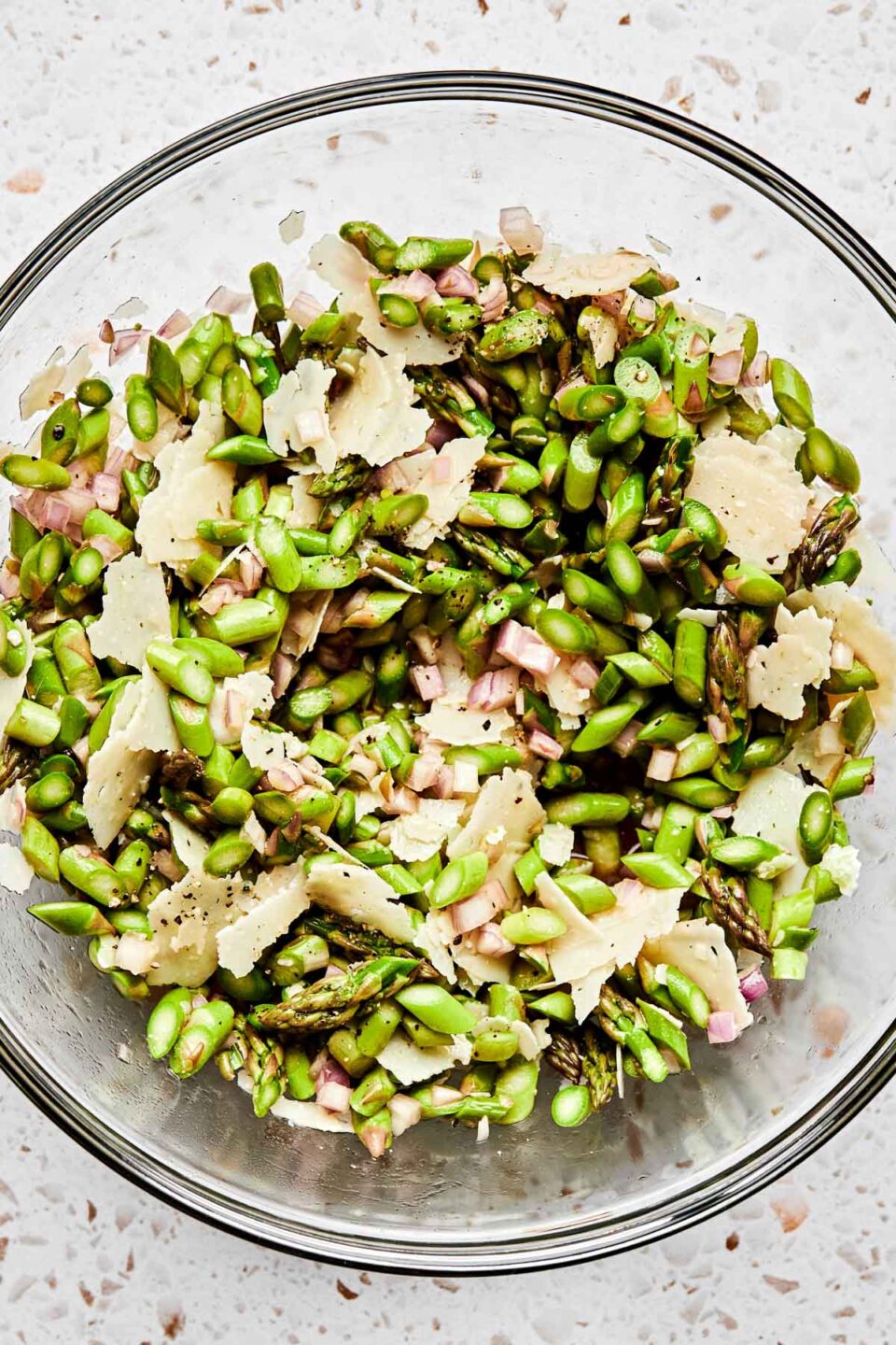 An overhead shot of raw asparagus salad in a large glass bowl atop a white textured surface.