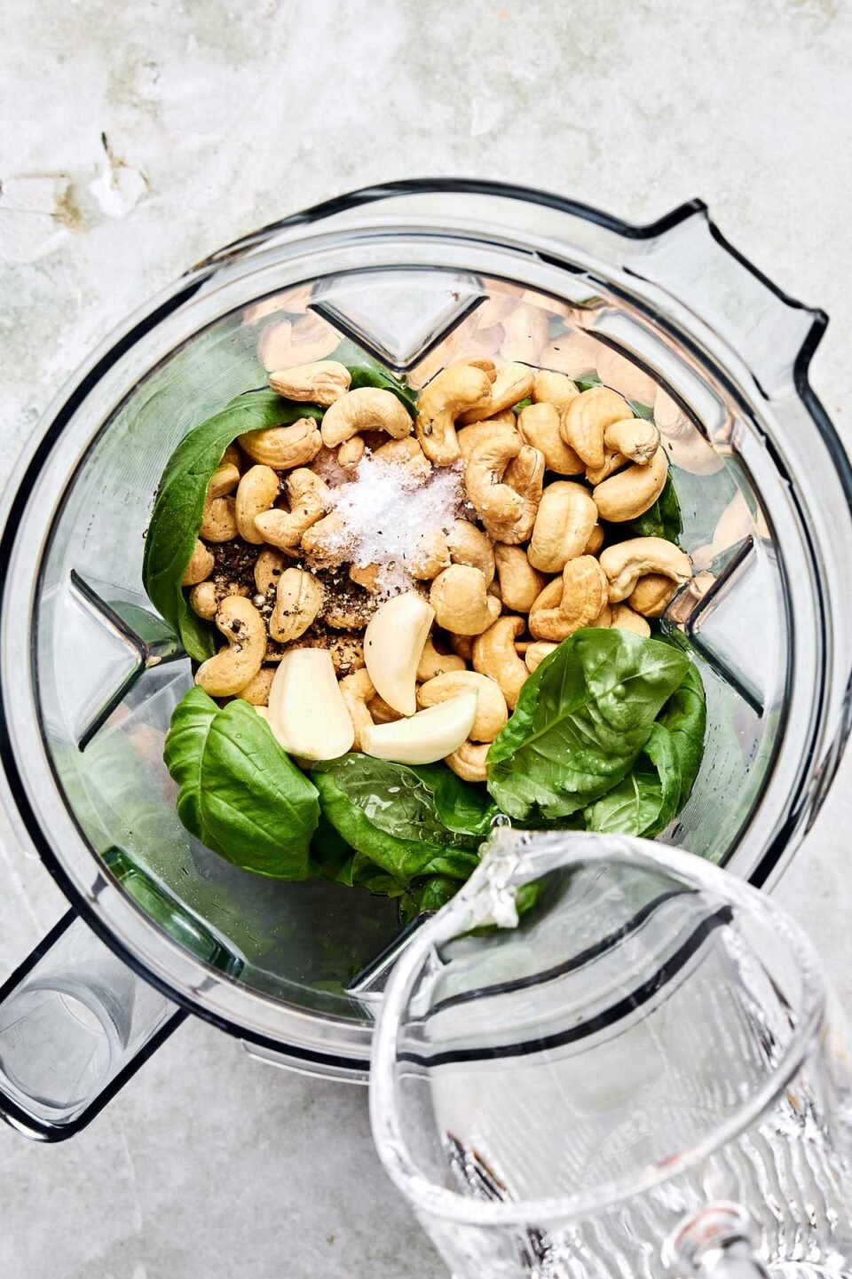 An overhead shot of water being poured into the bowl of a food processor containing cashews, basil, garlic, salt and pepper on a white textured surface.