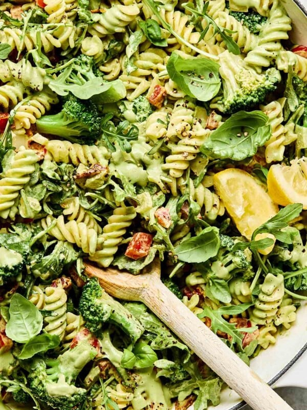 A close-up overhead shot of creamy vegan pasta in a white skillet, topped with basil leaves and lemon wedges.