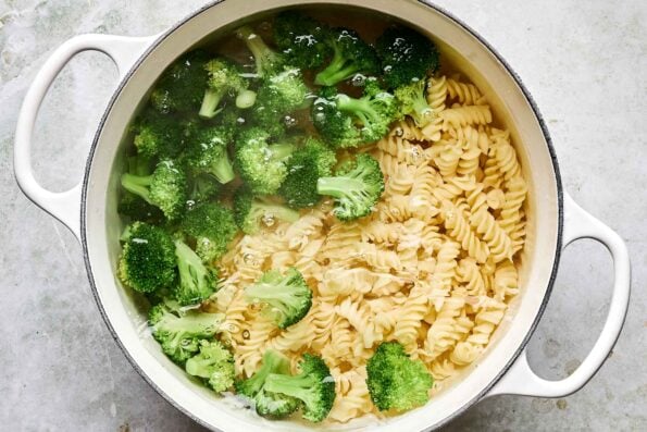 An overhead shot of pasta noodles and broccoli boiling in a large white pot upon a grey textured surface.