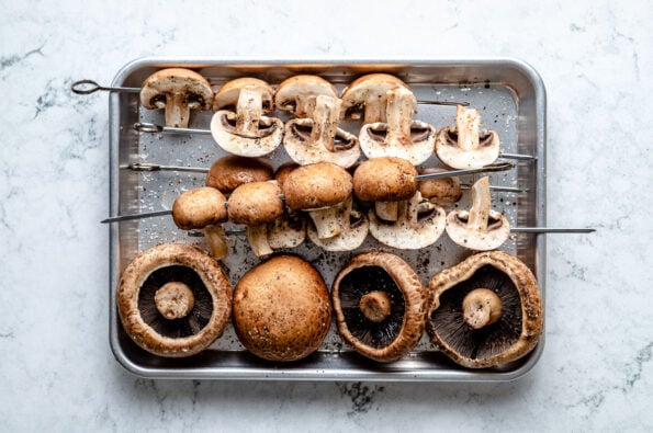 An overhead shot of prepared mushroom skewers and large whole mushrooms on a quarter sheet pan atop a white marbled surface.