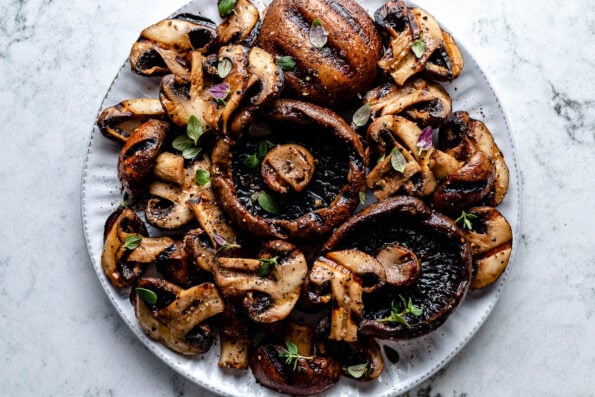 An overhead shot of grilled mushrooms of various types and sizes on a white plate atop a white marbled surface.