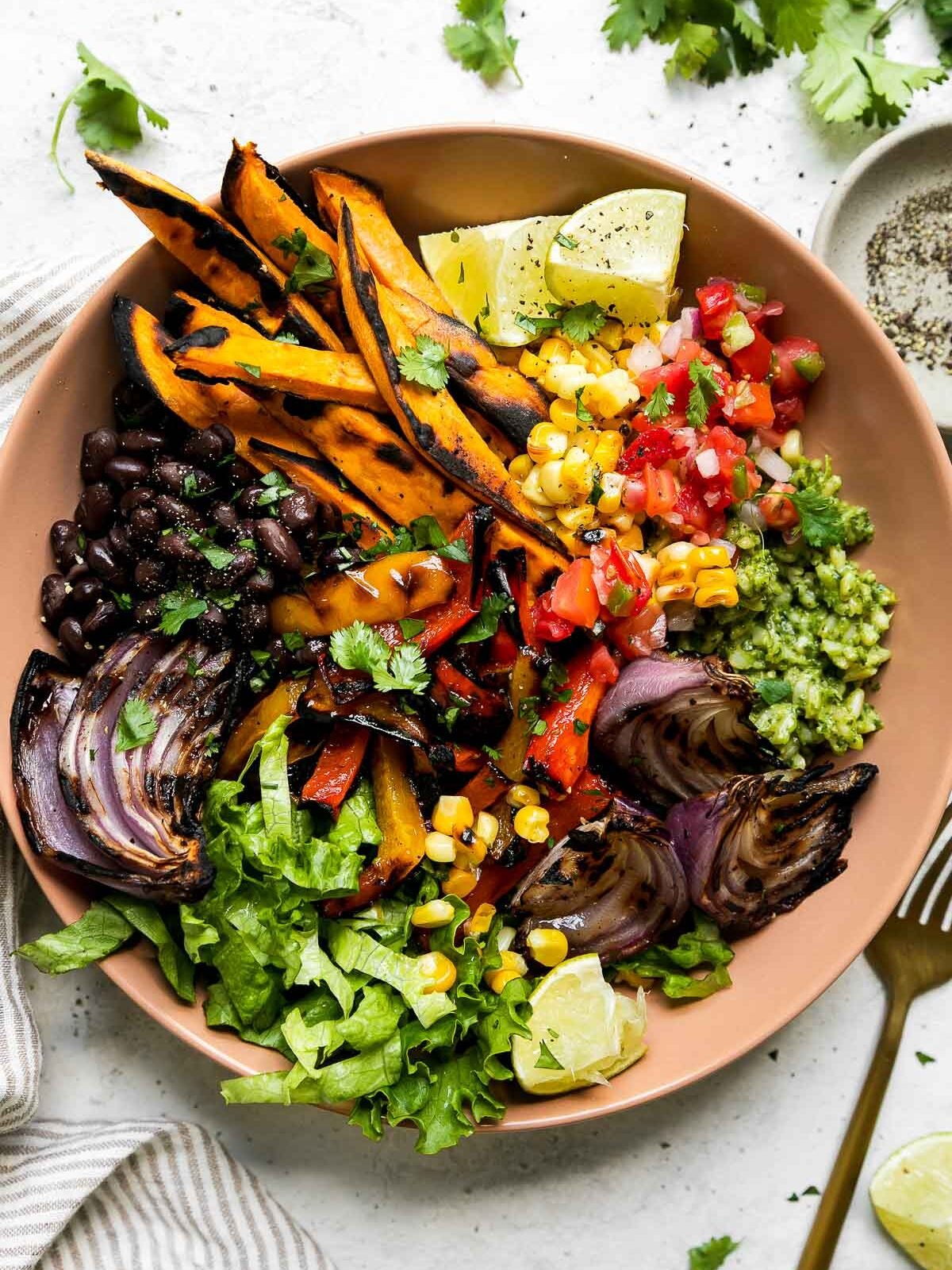 An overhead shot of a grilled vegetable burrito bowl inside of a pink tan ceramic bowl that sits atop a creamy white textured surface. A brown and white linen napkin, a small ceramic pinch bowl filled with ground black pepper, a gold fork, fresh cilantro, and a lime wedge surround the vegetarian burrito bowl at center.