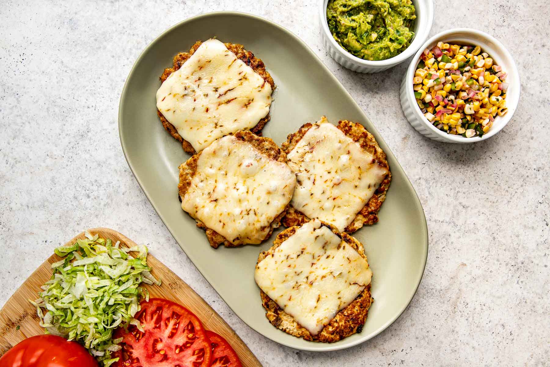 An overhead shot of four chicken smash burger patties topped with cheese on a light green oval platter atop a white textured surface. The platter is surrounded by dishes of guacamole and corn salsa and a cutting board with tomato slices and lettuce.