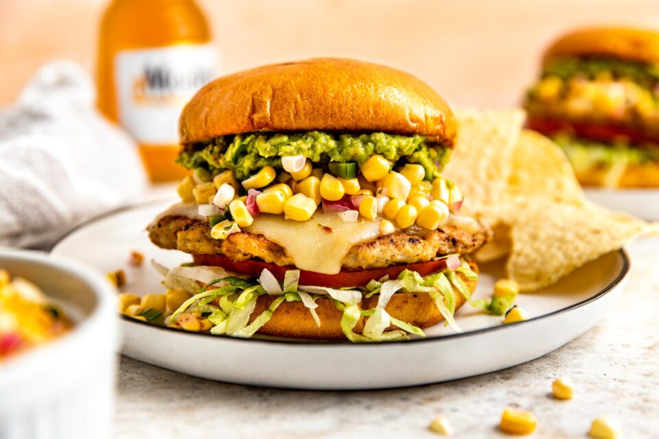 A side shot of a chicken smash burger topped with cheese, corn salsa and guacamole on a bun sitting on a white plate atop a white surface. A bottle of beer and a second burger sit in the background.
