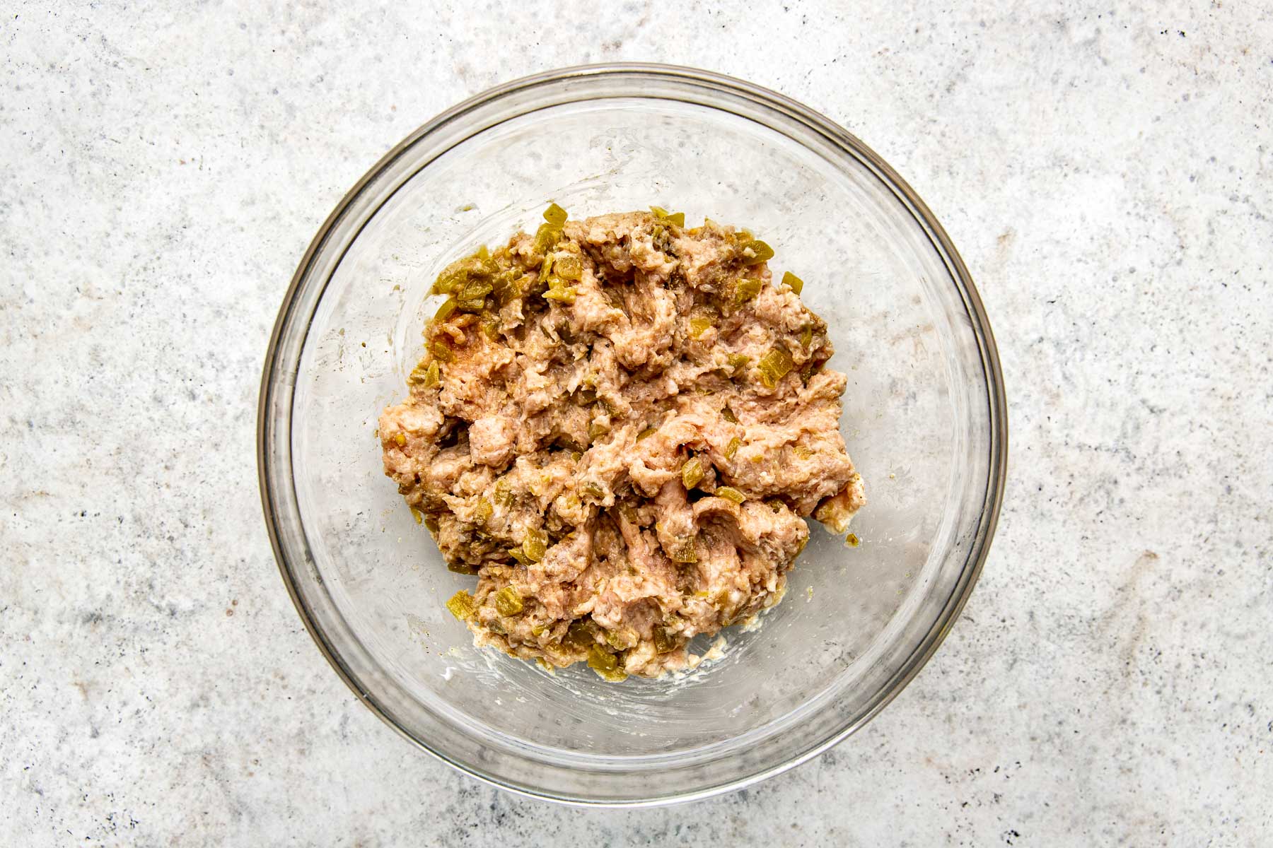 An overhead shot of the mixed burger mixture in a glass bowl atop a white textured surface.