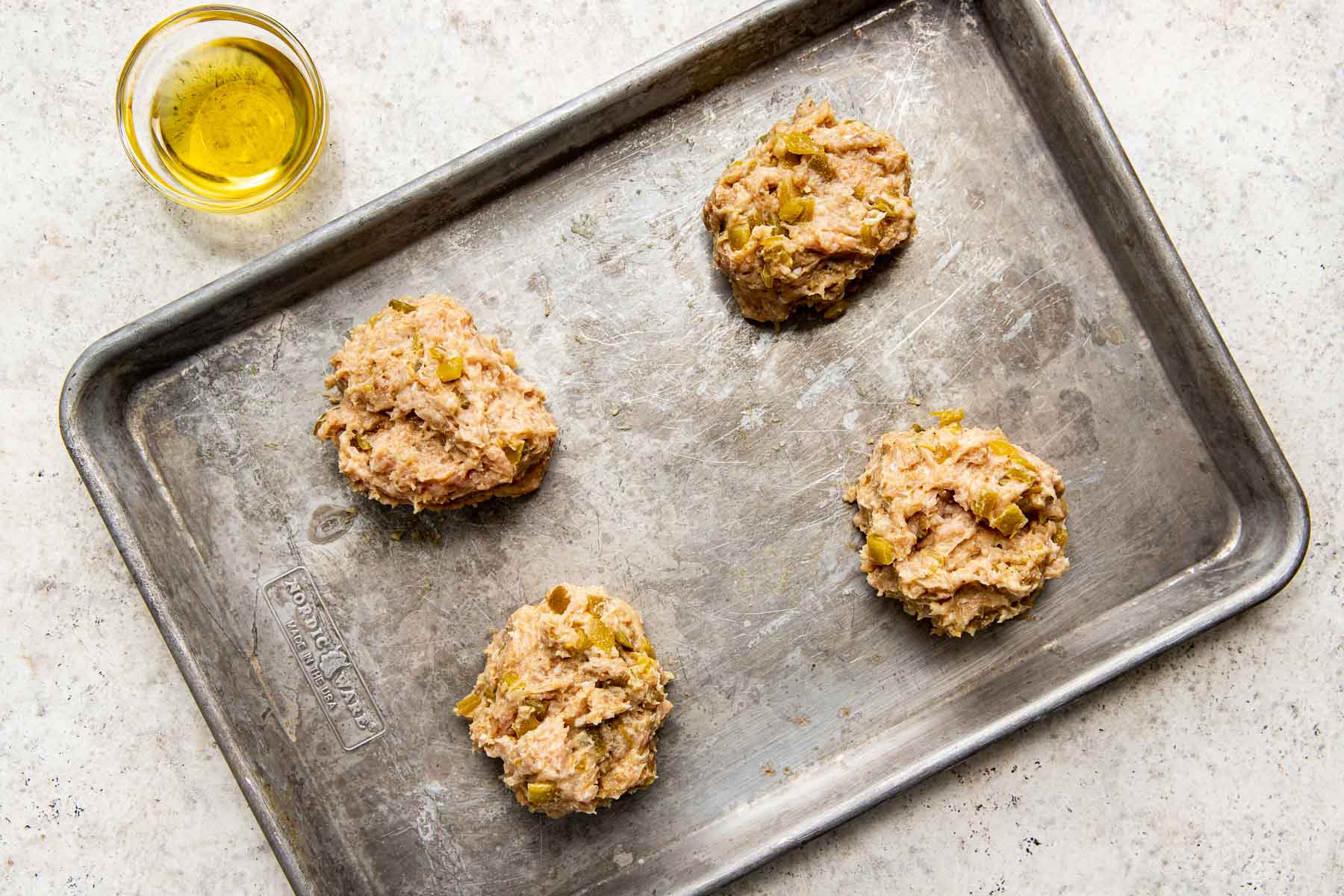 An overhead shot of four balls of burger mixture on a sheet pan atop a white textured surface alongside a bowl of oil.