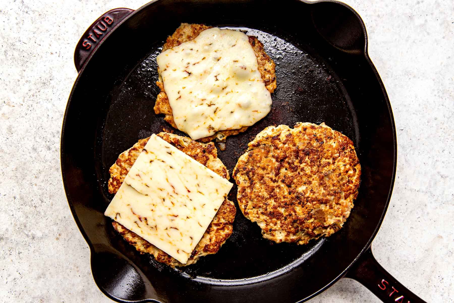Three burger patties, two with cheese and one without, in a black skillet atop a white textured surface.