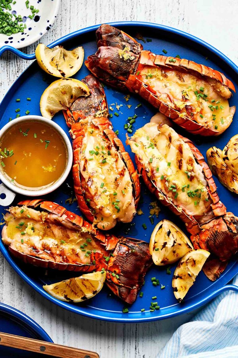 An overhead shot of grilled lobster tails garnished with chives on a bright blue plate. Lemon wedges and a small dish of melted butter sit alongside the tails.