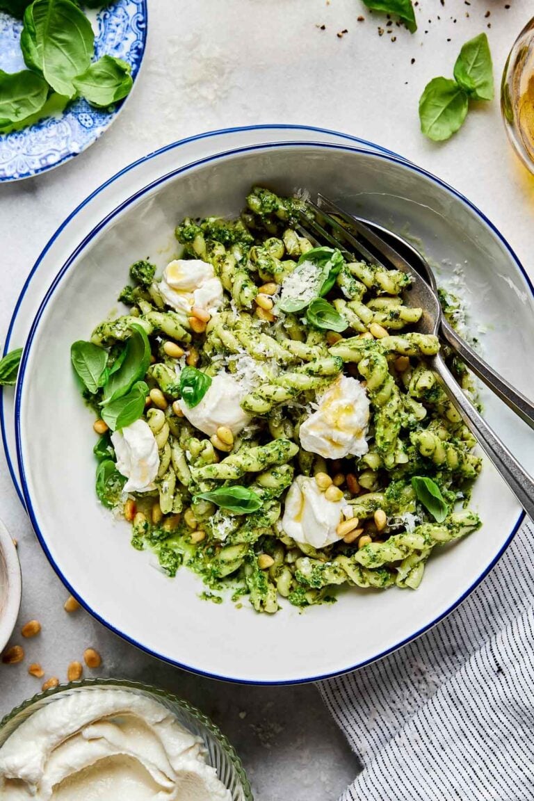 An overhead shot of pesto pasta topped with whipped ricotta and pine nuts in a large white bowl atop a white surface. A striped cloth and dishes of ricotta and fresh basil sit alongside the bowl.
