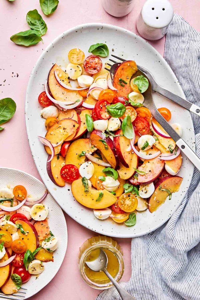 An overhead shot of a large white platter and a small white plate of tomato peach caprese salad on a light pink surface alongside a striped cloth and a jar of vinaigrette.