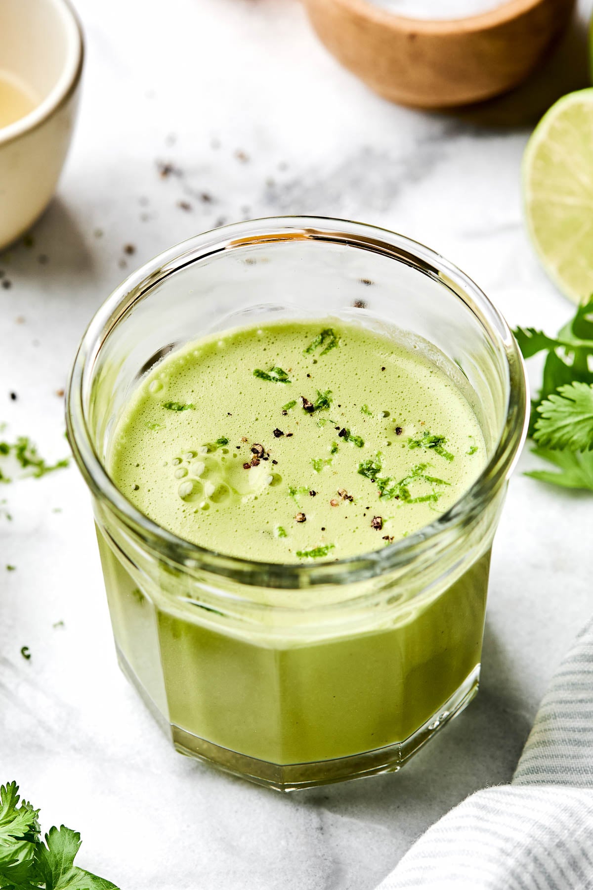 An angled shot of a clear glass of cilantro lime vinaigrette on a white marbled surface alongside cilantro leaves and a halved lime.