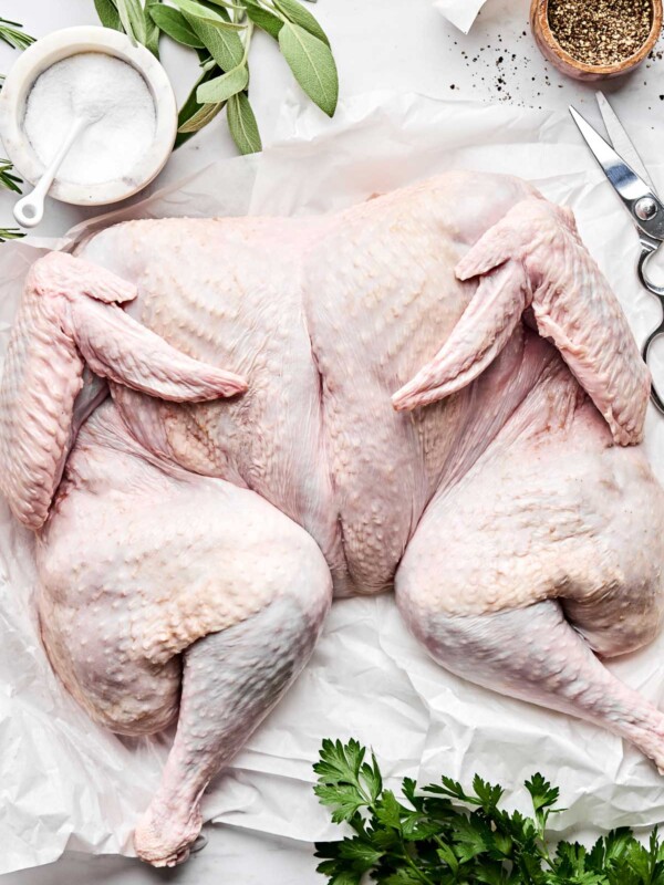 An overhead shot of a spatchcock raw turkey on a piece of parchment paper atop a white marbled surface. Fresh herbs, oil, garlic, butter, salt and pepper sit alongside the turkey.