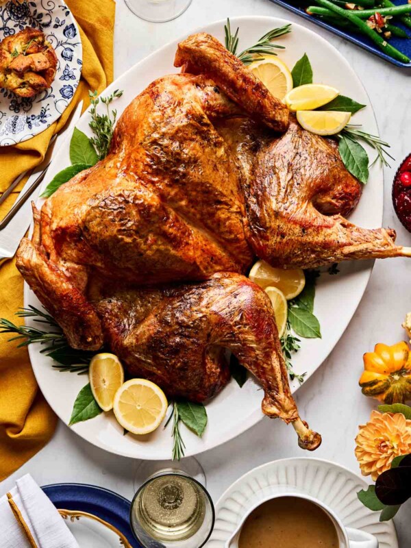 An overhead shot of a lemon herb roasted spatchcock turkey on a white oval platter garnished with herbs and lemons atop a white marble surface. The platter is surrounded by stuffing muffins, green beans, a bowl of gravy, a bowl of cranberries, and autumn flowers and gourds.