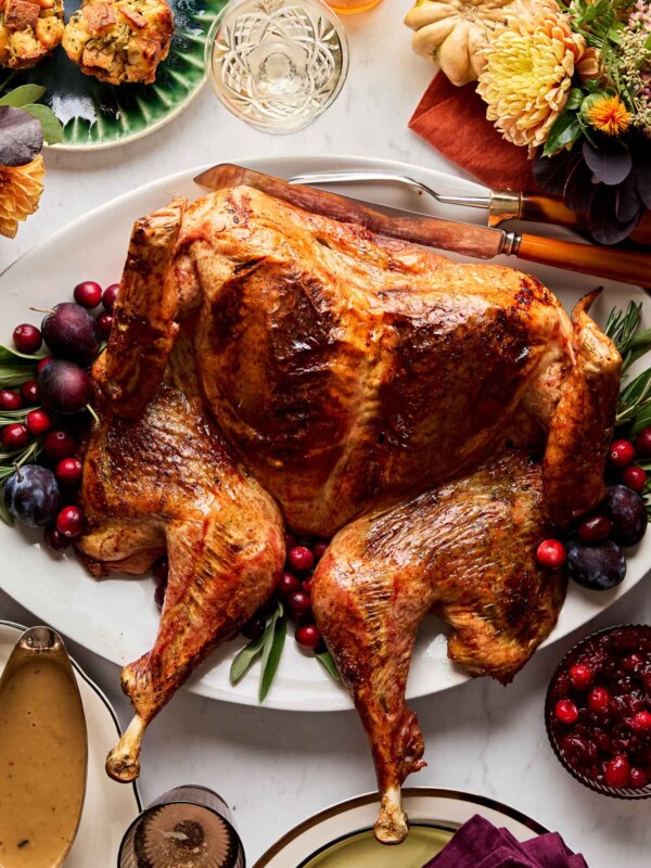 An overhead shot of a roasted maple glazed spatchcock turkey on a white oval platter garnished with herbs and cranberries atop a white marble surface. The platter is surrounded by stuffing muffins, green beans, a bowl of gravy, a bowl of cranberries, and autumn flowers and gourds.
