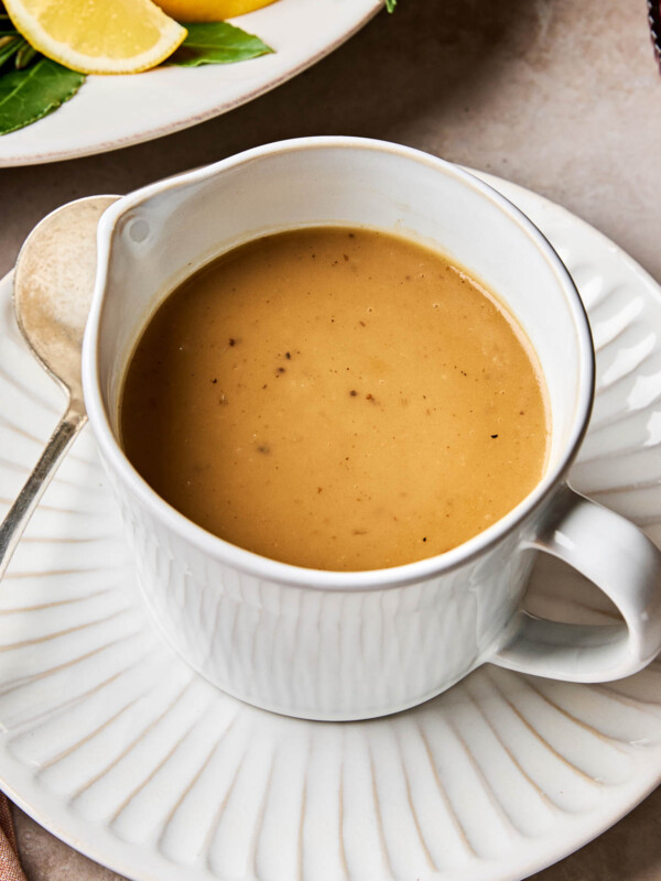A close-up angled shot of turkey gravy in a white gravy bowl atop a white scalloped plate. The plate sits on a beige surface.