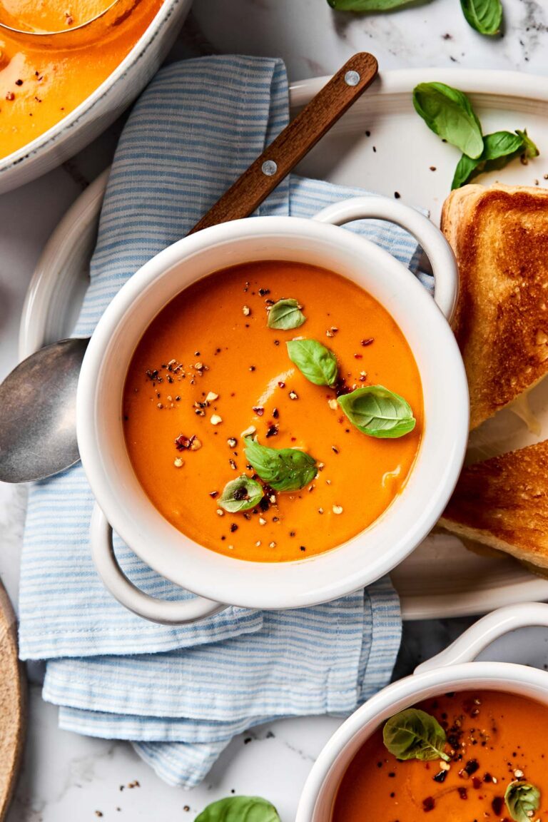An overhead shot of a small white handled bowl of creamy tomato soup topped with cracked pepper and basil leaves on a white marbled surface alongside a striped blue and white cloth. A second bowl of soup and a grilled cheese sandwich sit alongside it.