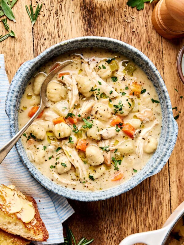 An overhead shot of a serving of chicken and gnocchi soup in a blue speckled bowl atop a wooden surface. The bowl is surrounded by fresh herbs, salt and pepper, and crusty bread on a blue striped cloth.