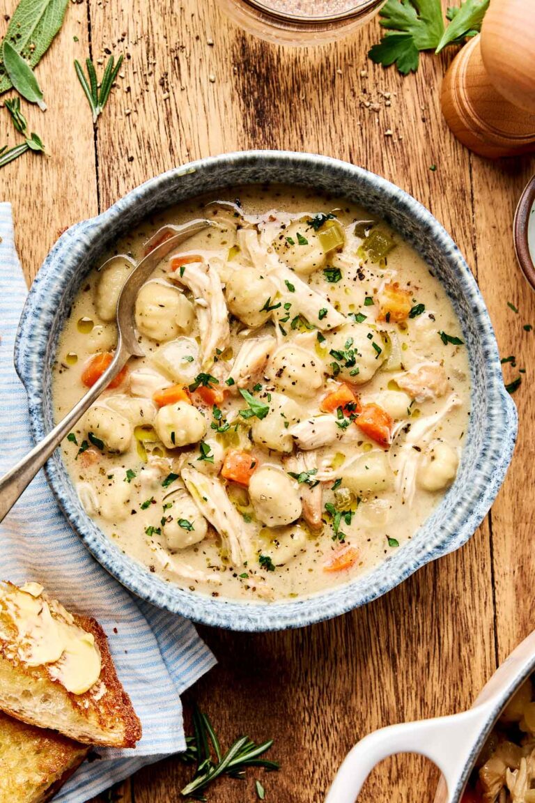 An overhead shot of a serving of chicken and gnocchi soup in a blue speckled bowl atop a wooden surface. The bowl is surrounded by fresh herbs, salt and pepper, and crusty bread on a blue striped cloth.