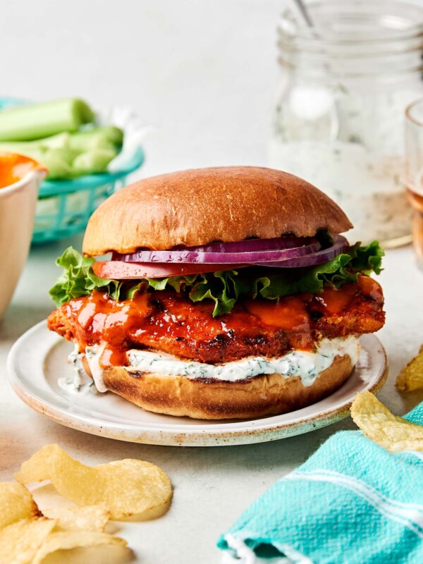 An side shot of an assembled buffalo chicken sandwich on a brioche bun atop a small white plate. The plate is surrounded by a bowl of buffalo sauce, a jar of ranch dressing, a basket of celery sticks, and potato chips on a white surface.