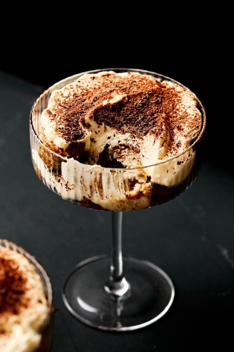 An angled shot of tiramisu in a coupe glass. A spoonful of the tiramisu has been removed from the glass.
