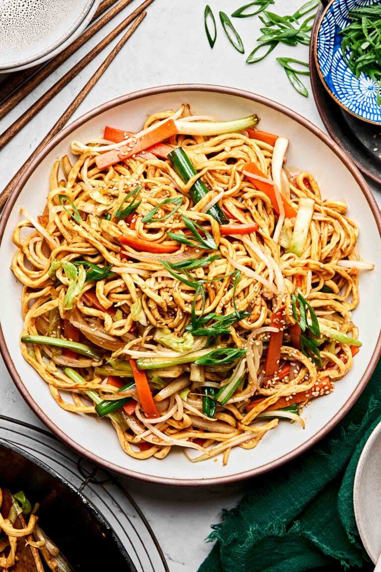 An overhead shot of stir fried vegetable noodles on a white stoneware plate on a white marbled surface. The plate is surrounded by a wok of noodles and a small blue dish of sliced green onions.